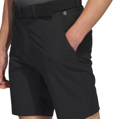 A person stands against a white background wearing Adidas Men's Ultimate365 8.5" Golf Shorts in black, featuring lightweight stretch fabric, a textured belt, subtle button detail, and their right hand in the pocket.