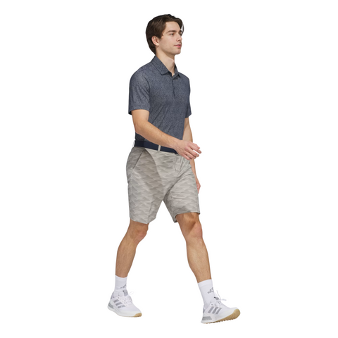 A young man is walking, wearing an Adidas Mens Ultimate365 Bunker Polo Shirt in Collegiate Navy, light gray patterned shorts, a navy belt, white socks, and white Adidas sneakers against a plain white background.