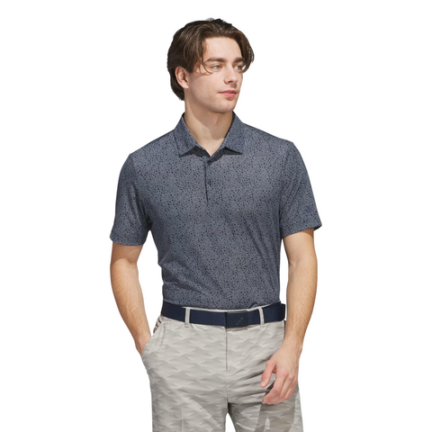 A man dressed in an Adidas Mens Ultimate365 Bunker Polo Shirt in Collegiate Navy, tucked into light gray patterned pants with a navy belt, stands with his hands in his pockets looking slightly to the side.