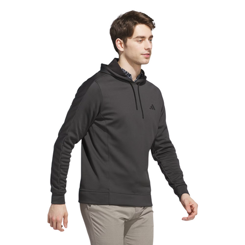 A man wears the Adidas Mens Ultimate365 Hoodie—a black fleece golf hoodie with four-way stretch and a small chest logo—paired with light beige pants, walking against a white background.