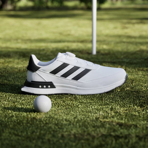 The Adidas Mens S2G BOA 24 Wide Spikeless Golf Shoe, in white with black stripes, rests on grass beside a golf ball, with a flag and green in the background.