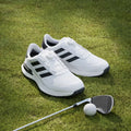 The Adidas Mens S2G BOA 24 Wide Spikeless Golf Shoes in white with black stripes sit on green grass beside a golf club and ball.