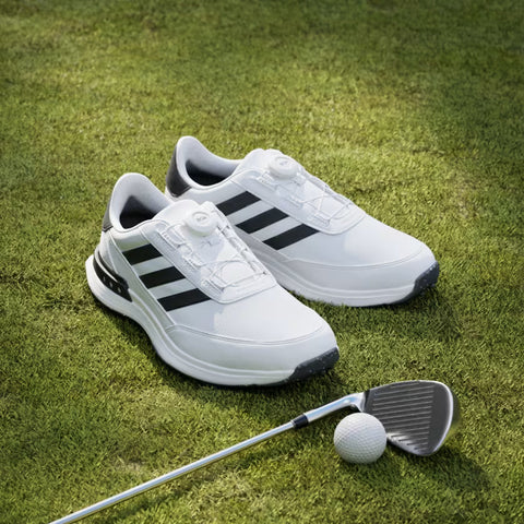 The Adidas Mens S2G BOA 24 Wide Spikeless Golf Shoes in white with black stripes sit on green grass beside a golf club and ball.