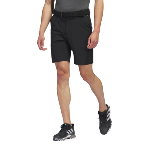 A man wearing Adidas Men's Ultimate365 8.5" Golf Shorts, a black belt, a dark fitted shirt, and black athletic shoes with white accents stands with one leg forward against a white background.