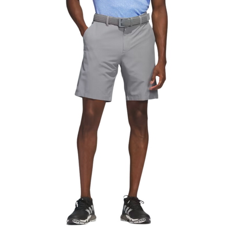 A man is shown from shoulders down wearing Adidas Ultimate365 8.5" Men's Golf Shorts in light gray, paired with a matching belt, black athletic shoes with white soles, and a light blue shirt on a white background, hands in pockets.