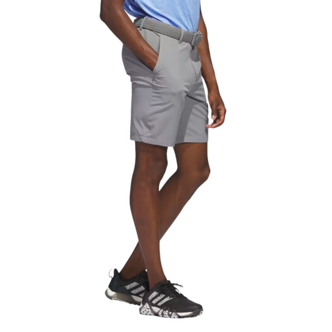 A person stands with hands in the pockets of Adidas Ultimate365 8.5" Men's Golf Shorts in grey, paired with a light blue shirt, black running shoes with white soles, and a gray belt. The image highlights their lower body.