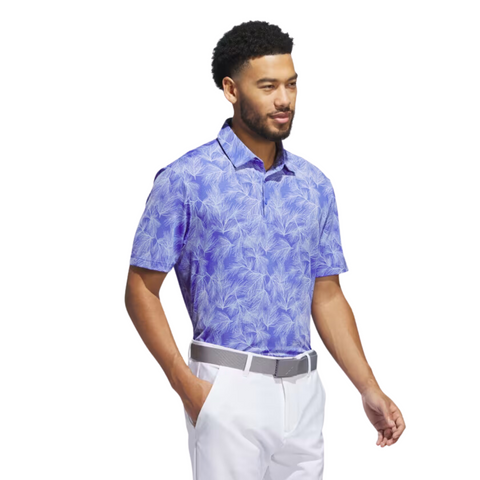 A man in a 2XL Adidas Ultimate365 Mesh Print Mens Polo by Adidas, featuring a blue and white palm leaf design, white pants, and a gray belt stands with one hand in his pocket against a white background.