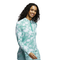 A woman with long curly hair smiles in a Pure Teal Adidas Ultimate365 Printed Quarter-Zip Long-Sleeve Polo Shirt and light pants, standing against a white background.