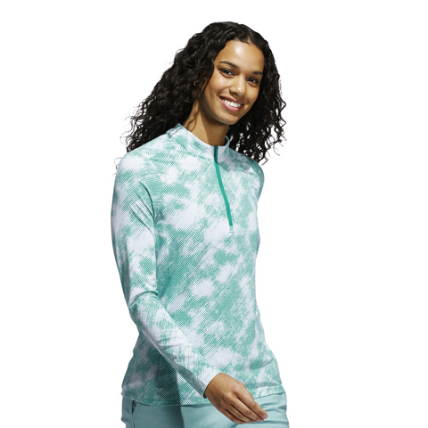 A woman with long curly hair smiles in a Pure Teal Adidas Ultimate365 Printed Quarter-Zip Long-Sleeve Polo Shirt and light pants, standing against a white background.