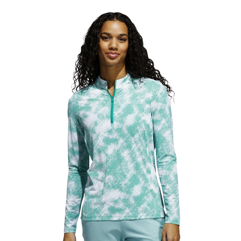 A woman with long curly hair wears an Adidas Ultimate365 Printed Quarter-Zip Long-Sleeve Polo Shirt in Pure Teal, paired with light blue pants, standing against a plain white background.