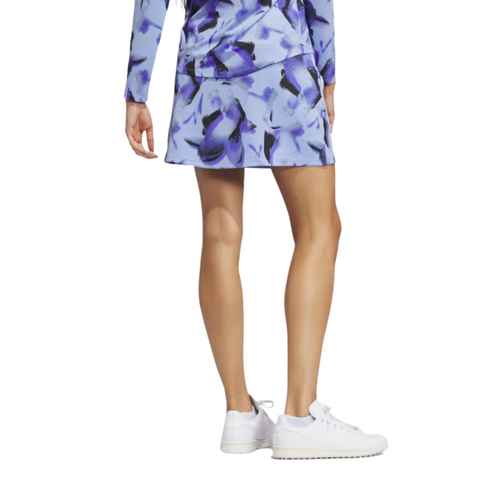 A woman is seen from the back wearing an Adidas Ultimate365 Women's Skort with a matching blue and purple abstract long-sleeve top and white sneakers.
