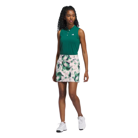 A woman in a sleeveless green polo, Adidas Ultimate365 Women's Skort by Adidas with a green leaf pattern, and white sneakers stands smiling against a white background.