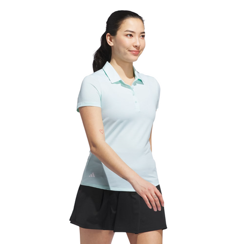 A smiling woman with dark hair in a ponytail wears the Adidas Women's Ottoman Short Sleeve Polo Shirt in Flash Aqua and black shorts, walking forward against a white background.
