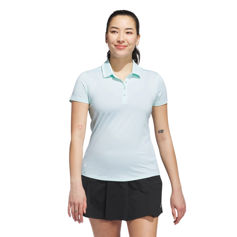 A woman with tattoos on her arms, including a heart outline on her left arm, stands smiling against a white background wearing an Adidas Women's Ottoman Short Sleeve Polo Shirt in Flash Aqua and a black skirt.
