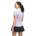 A woman with a ponytail, wearing an Adidas Women’s Ottoman Short Sleeve Polo Shirt in Powder Plum and a black pleated skirt, stands facing away and smiles over her shoulder.