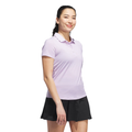A woman with her hair tied back and a small heart tattoo on her left arm smiles in an Adidas Women’s Ottoman Short Sleeve Polo Shirt in Powder Plum, paired with a black skirt, against a white background.