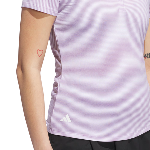 A person wears an Adidas Women’s Ottoman Short Sleeve Polo Shirt in Powder Plum with black pants. A small red heart outline tattoo is visible on their inner arm.
