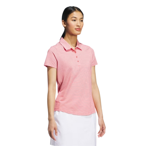 A woman with long dark hair pulled back poses against a white background wearing an Adidas Women's Ottoman Short Sleeve Polo Shirt in Pure Ruby and a white skirt.