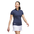 A woman wearing an Adidas Womens Ultimate365 Jacquard Short Sleeve Polo Shirt in Collegiate Navy and a white skirt stands against a white background, showing small arm tattoos as she smiles and looks to the side.