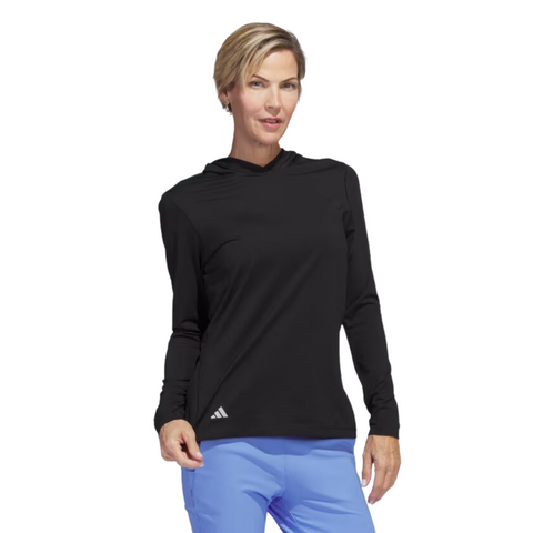 Model wearing Adidas women's performance golf hoodie in black. The hoodie has a sleek, moisture-wicking fabric, long sleeves, and a white Adidas logo on the lower left side. The model pairs it with light blue golf pants.