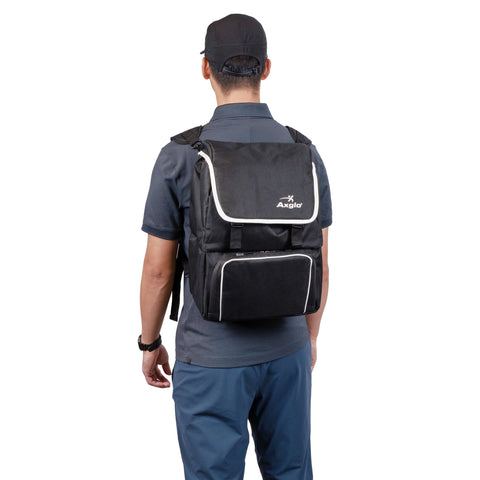 A person is seen from behind wearing a black cap, dark gray shirt, and blue pants, carrying a large black Angio backpack with white trim and an Axglo Cooler Bag by Axglo attached.