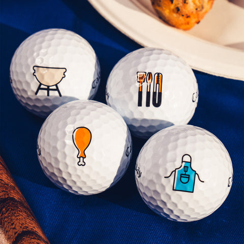 Four white Callaway 2025 Supersoft Grill Master Golf Balls by Callaway sit on a blue cloth, each featuring a unique barbecue-themed icon: grill, utensils with thermometer, chicken drumstick, and apron for ultimate soft feel.
