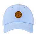 The Callaway Women’s Heritage Twill Golf Cap in light blue features a brown circular leather patch with the Callaway name and black chevron logo, plus an adjustable fit for stylish sun protection.