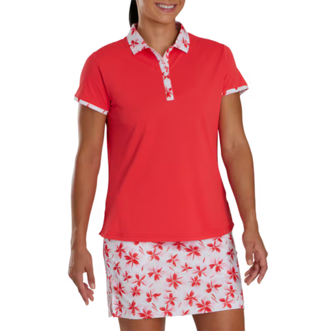 A woman wears the FootJoy Floral Trim Short Sleeve Womens Polo, a coral moisture-wicking golf shirt with floral trim, paired with a white skirt featuring a coordinating coral and red flower pattern.