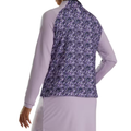 A woman is seen from the back wearing a light purple FootJoy Full-Zip Printed Pullover, paired with a darker purple and black patterned vest. Her left hand rests on her hip, and she completes the look with a matching skirt.