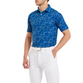 A man wears the FootJoy Golf Course Doodle Mens Polo, a blue patterned, moisture-wicking shirt from FootJoy, paired with white pants and a white belt, standing with one hand in his pocket against a plain background.