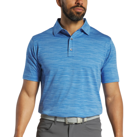 A man wears a FootJoy Jacquard Wave Mens Polo in light blue with a subtle wave pattern, paired with gray pants and a light gray belt, standing against a plain white background.