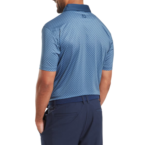 A man is seen from the back wearing a FootJoy Men's Checker Print Lisle Polo in Dark Denim/White, paired with navy pants and a matching belt. His hands are in his pockets, and he looks slightly to the side.