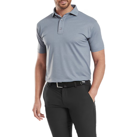 A man wears the FootJoy Men's Etched Texture Print Lisle Polo in Flint, short-sleeved and tucked into black pants with a belt, standing with one hand in his pocket against a white background.
