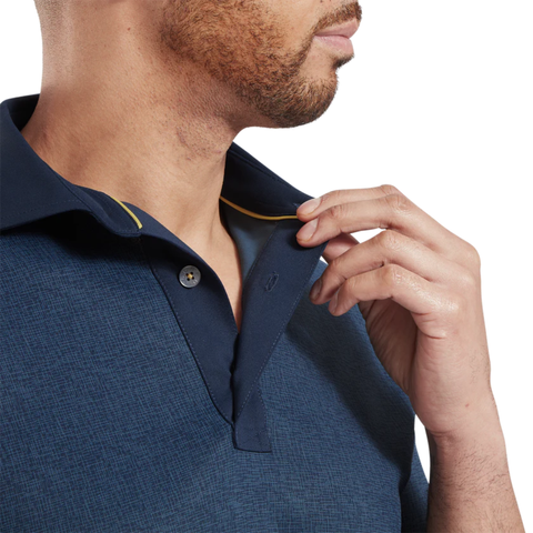 A man in a FootJoy Men's Etched Texture Print Lisle Polo in navy gently holds his collar, revealing a yellow trim. The cropped image focuses on the lower half of his face and upper torso, showcasing this stylish FootJoy golf shirt.