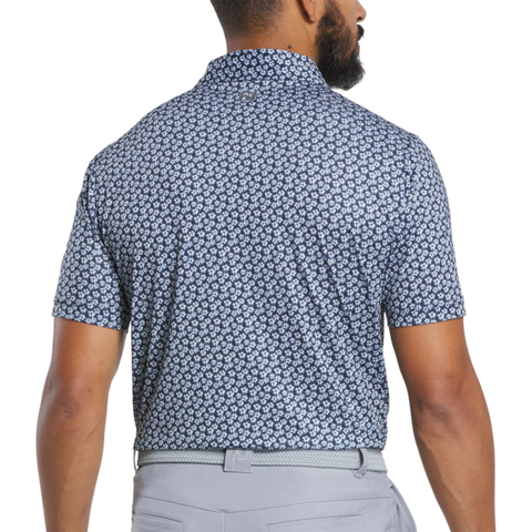 A man faces away wearing the FootJoy Men's Floral Sketch Print Lisle Polo in navy and white, tucked into light gray pants with a matching belt.