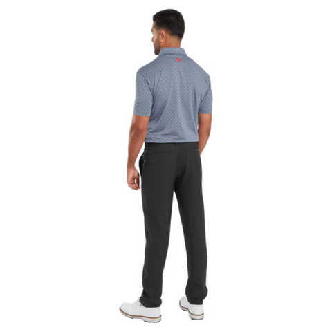 A man faces away from the camera wearing a blue FootJoy Men's Flower Bud Foulard Lisle Polo in Flint, featuring moisture-wicking fabric, paired with black pants and white shoes, on a plain white background.