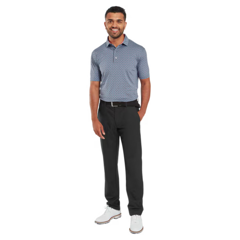 A man smiles in a blue FootJoy Men's Flower Bud Foulard Lisle Polo | Flint, styled with black pants, a black belt, and white golf shoes. He stands against a plain white background.