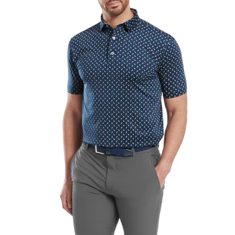 A man wears the FootJoy Men's Flower Bud Foulard Lisle Polo in navy, tucked into gray pants with a navy belt. One hand is in his pocket and his head is turned slightly to the side.