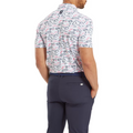 A man stands with his hands in his pockets, wearing navy pants and a FootJoy Men’s Golf Course Doodle Polo by FootJoy, featuring a white performance stretch fabric with an abstract pink, blue, and black pattern. The image highlights the polo's unique back design.