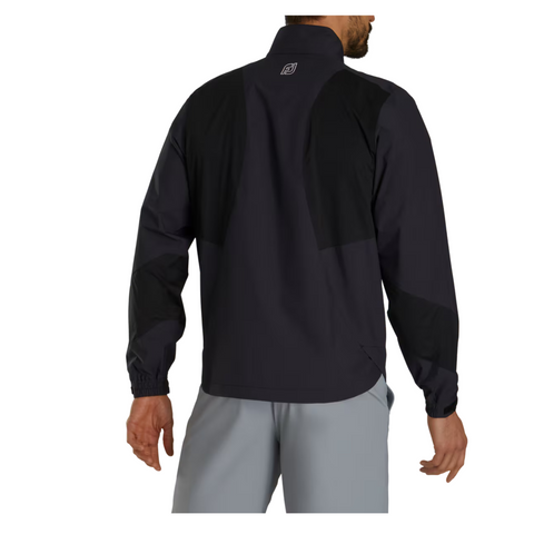 A man is seen from the back wearing a FootJoy Men’s HydroLite X Rain Jacket in black with a high collar, black panel details, and elastic-cuffed long sleeves, paired with light gray pants. This waterproof golf jacket is made by FootJoy.