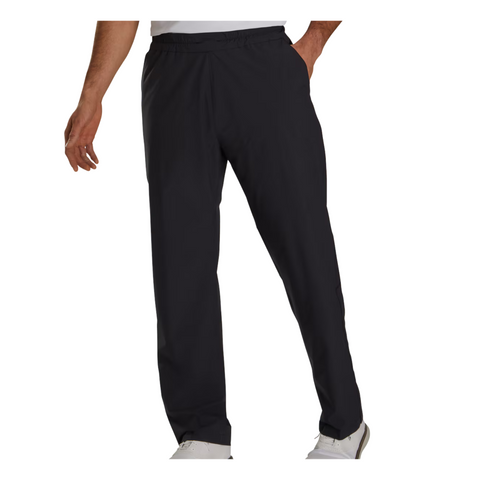 A man wearing FootJoy Men’s HydroLite X Rain Pants in black, paired with a white shirt and white shoes, stands with one hand by his side and the other on his hip.