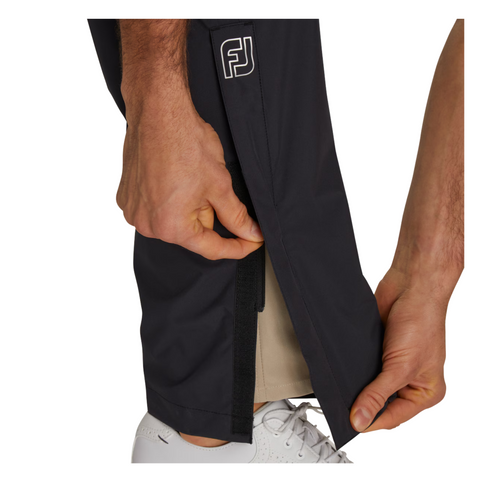 A person in black FootJoy Men’s HydroLite X Rain Pants with an FJ logo opens the Velcro ankle flap to reveal beige fabric underneath. They are also wearing a white shoe.