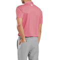 A man, seen from behind, wears a FootJoy Men's Lisle Houndstooth Geo Print Polo in Hibiscus and light gray pants, his left hand in his pocket.