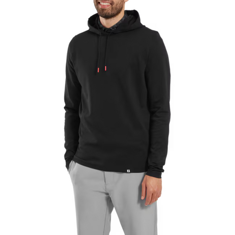 A man wearing a FootJoy Men's Ottoman Jacquard Pullover Hoodie in black and light gray pants stands with one hand in his pocket, smiling against a plain white background.