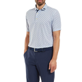 A man wears a FootJoy Mens Pique Solid Figure Print Lisle Polo in White/Navy, tucked into navy pants with a matching belt. His head is slightly turned and his left hand is in his pocket; the polo’s moisture-wicking fabric adds comfort.