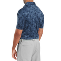 A man is seen from the back wearing a FootJoy Men's Pop Camo Lisle Polo in Dark Denim and light gray pants with a textured belt. His relaxed pose highlights the stylish fit and moisture-wicking comfort of this FootJoy golf polo.
