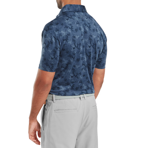 A man is seen from the back wearing a FootJoy Men's Pop Camo Lisle Polo in Dark Denim and light gray pants with a textured belt. His relaxed pose highlights the stylish fit and moisture-wicking comfort of this FootJoy golf polo.