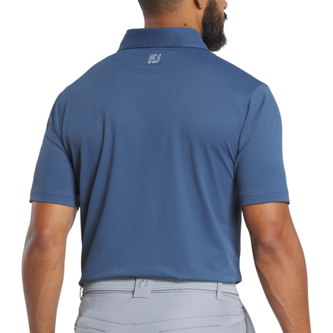 A man with a short beard is seen from behind wearing FootJoy Men's Solid Pique with Trim Polo in Dark Denim/Skyway, featuring an FJ logo on the back collar, paired with light gray pants. His right arm is slightly bent.