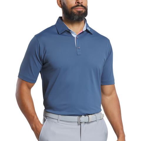 A man wears a FootJoy Men's Solid Pique with Trim Polo in Dark Denim/Skyway, paired with light gray pants and a matching belt, standing against a plain white background.
