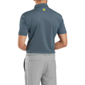 A man wearing a FootJoy Men’s Wiggle Print Lisle Polo in navy/white and light gray pants stands with his back to the camera. The polo features ProDry Performance Fabric and a small yellow logo at the upper back.